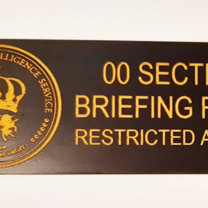 May include: A black oval sign with gold lettering that reads "00 Section Briefing Room Restricted Access". The sign also features a gold emblem with a crown and the words "Royal Secret Intelligence Service".
