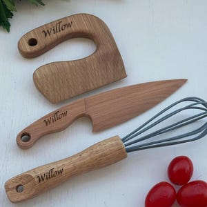 Puede incluir: Juego de utensilios de cocina de madera, que incluye un cortador curvo, un cuchillo y un batidor, con el nombre "Willow" grabado en cada uno. El conjunto se complementa con tomates cherry rojos, lo que sugiere un tema culinario.