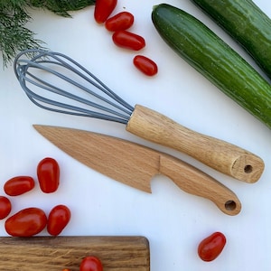 Puede incluir: Una tabla de cortar de madera con un cuchillo de madera y un batidor. Hay tomates cherry rojos y dos pepinos verdes sobre una superficie blanca.