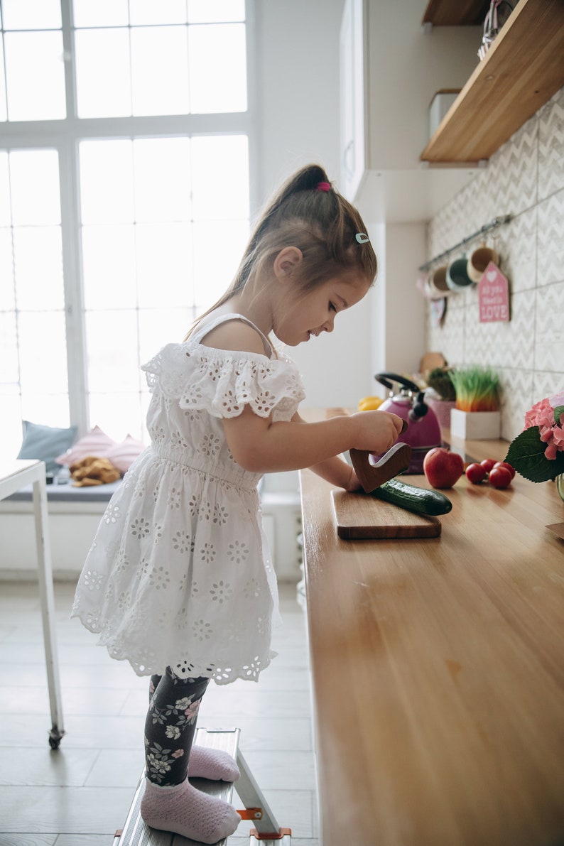 May include: A young girl in a white dress with floral embroidery stands on a step stool in a kitchen. She is using a wooden knife to cut a cucumber on a wooden cutting board. There are other vegetables on the counter, including tomatoes and green onions.