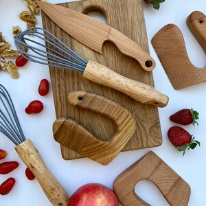 Puede incluir: Un conjunto de herramientas de juego de cocina de madera para ni&ntilde;os, que incluye una tabla de cortar, un cuchillo, un batidor y dos formas de esp&aacute;tula. Las herramientas est&aacute;n dispuestas sobre una superficie blanca con manzanas rojas, fresas y tomates.