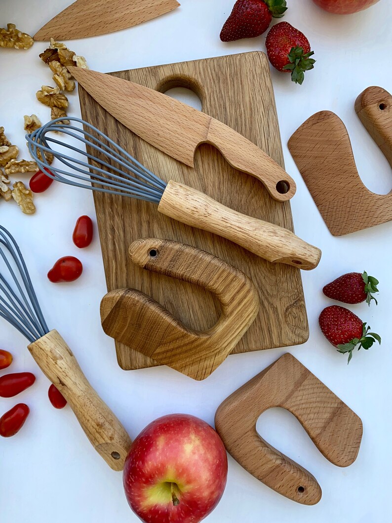 Puede incluir: Un conjunto de herramientas de juego de cocina de madera para ni&ntilde;os, que incluye una tabla de cortar, un cuchillo, un batidor y dos formas de esp&aacute;tula. Las herramientas est&aacute;n dispuestas sobre una superficie blanca con manzanas rojas, fresas y tomates.