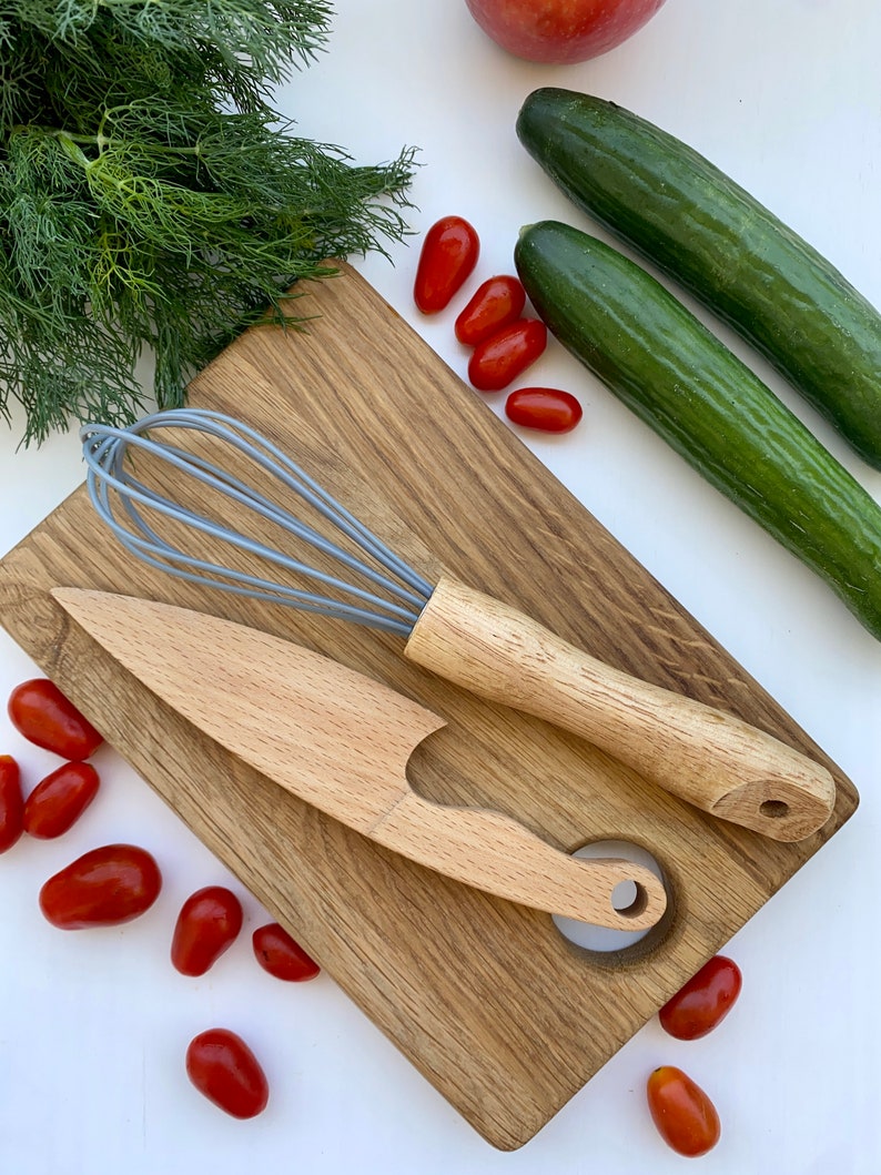 Puede incluir: Una tabla de cortar de madera con un batidor, un cuchillo de madera y un agujero en el centro. Hay tomates rojos y pepinos verdes alrededor de la tabla.