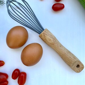 Puede incluir: Un batidor gris y de madera con huevos marrones y tomates rojos sobre una superficie blanca.