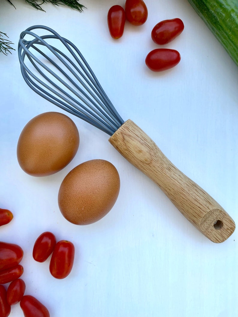 Puede incluir: Un batidor gris y de madera con huevos marrones y tomates rojos sobre una superficie blanca.