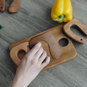 May include: A wooden serving board with three carved wooden handles. The board has a round cutout in the center and a smaller cutout on the left side. A hand is holding one of the handles.