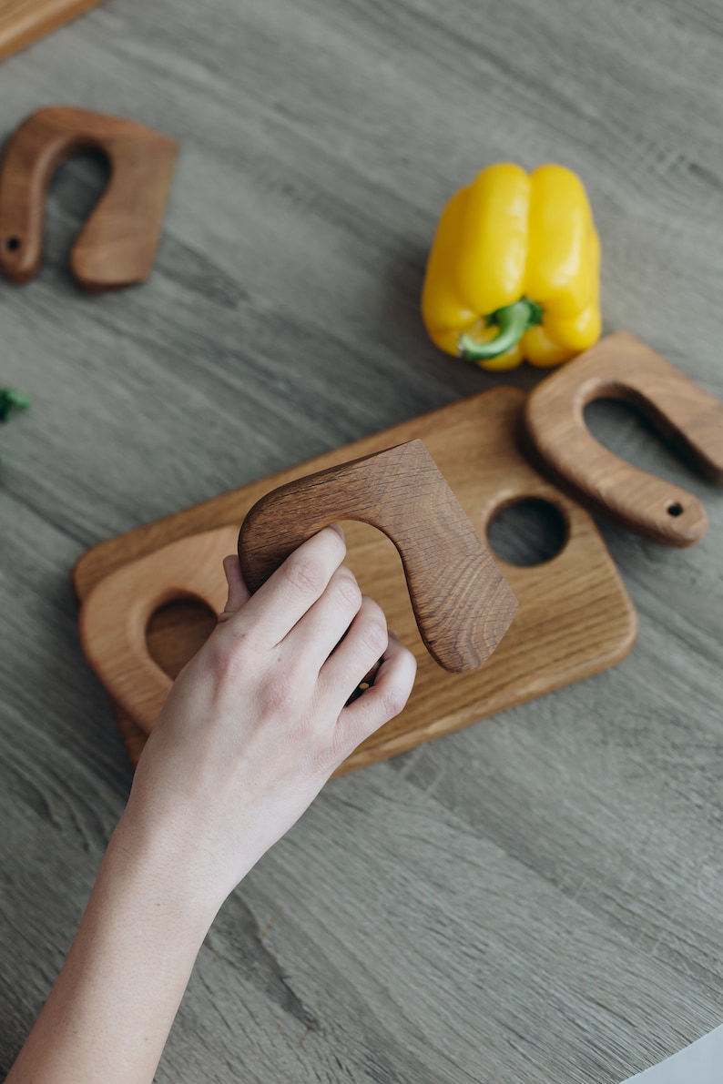 May include: A wooden serving board with three carved wooden handles. The board has a round cutout in the center and a smaller cutout on the left side. A hand is holding one of the handles.