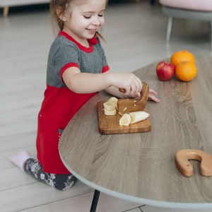 May include: A young girl in a red and grey dress is sitting at a wooden table, using a wooden knife to cut a banana into slices. There are other pieces of fruit on the table, including a red apple and two oranges.