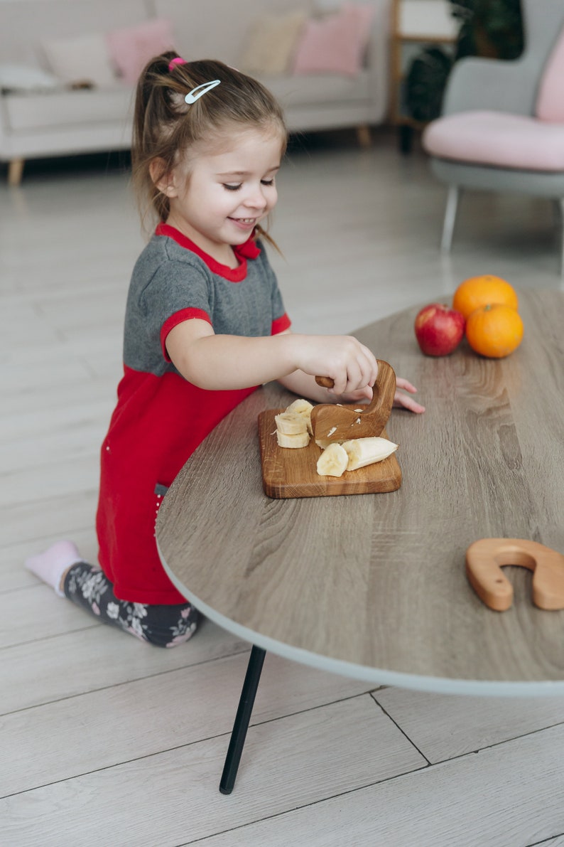 May include: A young girl in a red and grey dress is sitting at a wooden table, using a wooden knife to cut a banana into slices. There are other pieces of fruit on the table, including a red apple and two oranges.