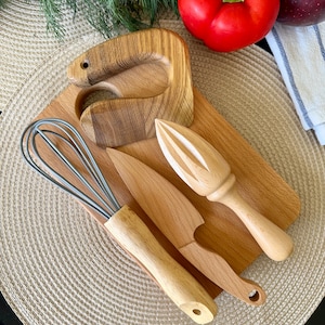 Op de afbeelding: Een set houten keukengereedschap op een houten snijplank. De set bevat een garde, een citruspers, een mes en een houten handvat.