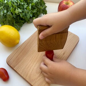 May include: A child's hand uses a wooden mallet to crush a red tomato on a wooden cutting board. There are other tomatoes and a lemon on the white surface.