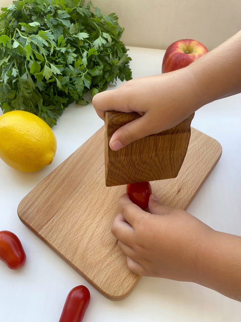 May include: A child's hand uses a wooden mallet to crush a red tomato on a wooden cutting board. There are other tomatoes and a lemon on the white surface.