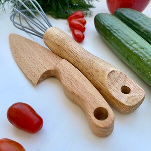 Puede incluir: Un juego de utensilios de cocina de madera para jugar, que incluye un cuchillo, una esp&aacute;tula y un batidor. Los utensilios est&aacute;n hechos de madera natural y tienen un color marr&oacute;n claro. Los utensilios est&aacute;n dispuestos sobre una superficie blanca con algunos tomates rojos y pepinos verdes.