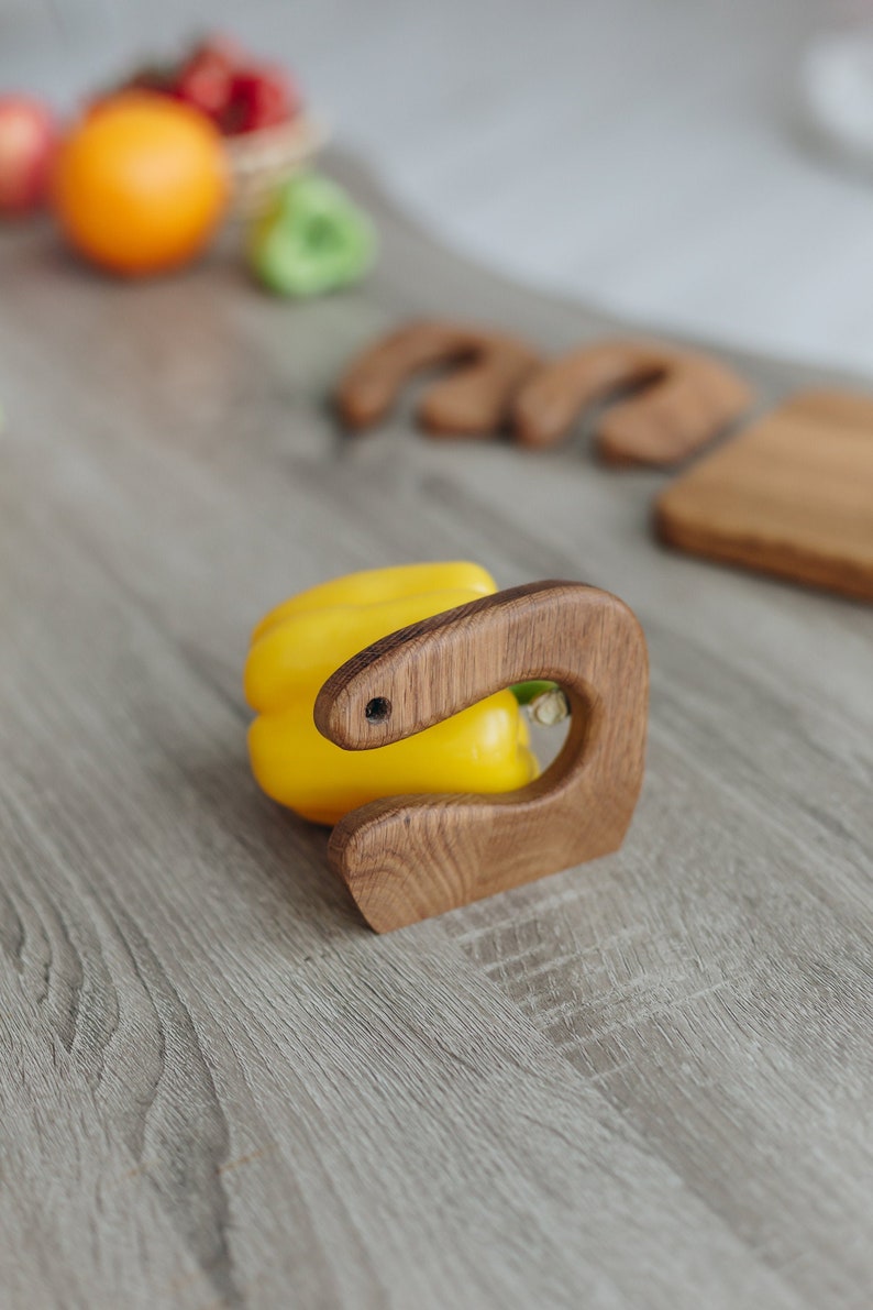 May include: A wooden kitchen utensil with a curved handle, designed to hold a yellow bell pepper. The utensil is made of natural wood and has a rustic finish.