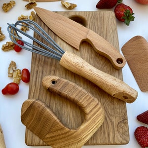 Puede incluir: Un juego de utensilios de cocina de madera en una tabla de cortar de madera. El juego incluye un batidor, dos cuchillos, una esp&aacute;tula y un mango. Las herramientas est&aacute;n hechas de madera natural y tienen un aspecto r&uacute;stico.