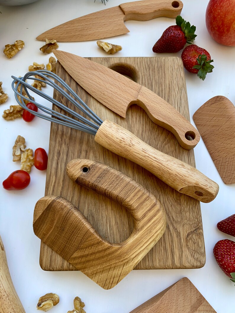 Puede incluir: Un juego de utensilios de cocina de madera en una tabla de cortar de madera. El juego incluye un batidor, dos cuchillos, una esp&aacute;tula y un mango. Las herramientas est&aacute;n hechas de madera natural y tienen un aspecto r&uacute;stico.