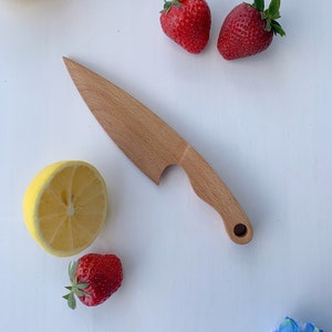 Puede incluir: Un cuchillo de madera con mango y hoja redondeada. El cuchillo est&aacute; sobre una superficie blanca con dos fresas rojas, un lim&oacute;n y medio lim&oacute;n.