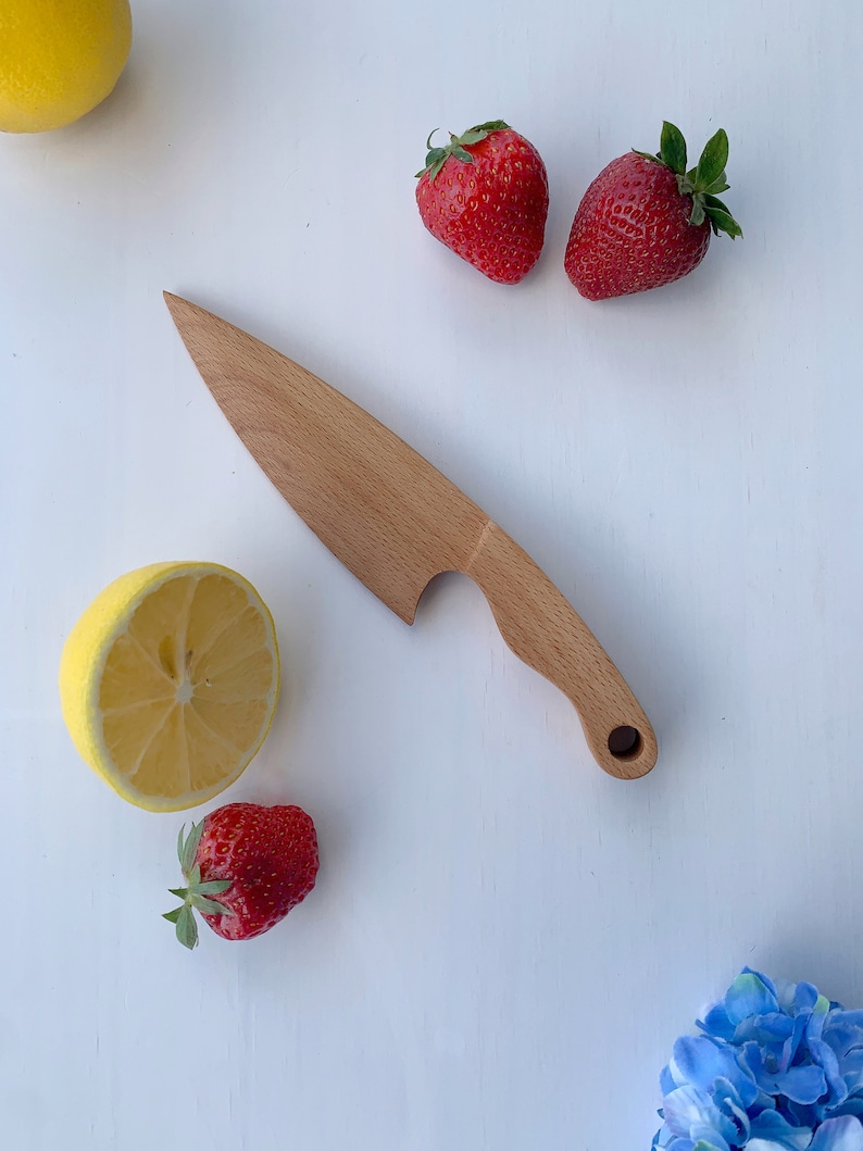 Puede incluir: Un cuchillo de madera con mango y hoja redondeada. El cuchillo est&aacute; sobre una superficie blanca con dos fresas rojas, un lim&oacute;n y medio lim&oacute;n.