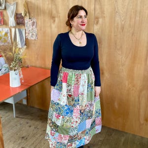 Puede incluir: Una mujer con una camisa negra de manga larga y una falda de retazos con estampados florales en tonos rosa, azul, verde y blanco. La falda está hecha de diferentes telas y tiene una cintura fruncida.