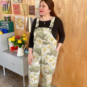 May include: A person wearing a pair of beige overalls with a dandelion print. The overalls have adjustable straps and pockets. The person is standing in front of a wooden wall.