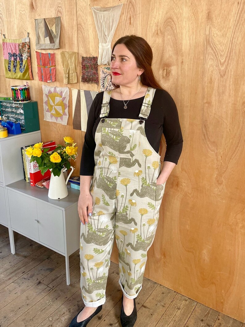 May include: A person wearing a pair of beige overalls with a dandelion print. The overalls have adjustable straps and pockets. The person is standing in front of a wooden wall.