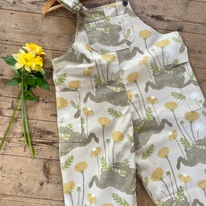May include: Beige overalls with a pattern of brown hares and yellow dandelions. The overalls have adjustable straps and a single front pocket.