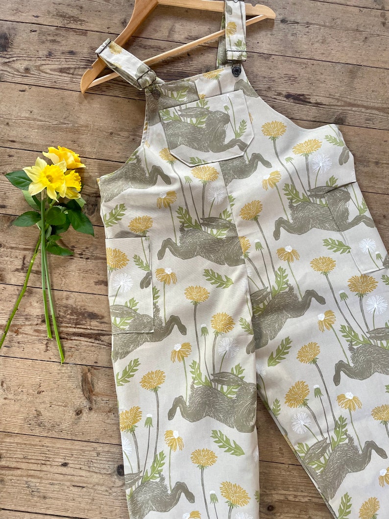 May include: Beige overalls with a pattern of brown hares and yellow dandelions. The overalls have adjustable straps and a single front pocket.