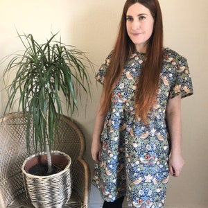May include: A woman wearing a blue and green floral dress with short sleeves. The dress has a pattern of birds and flowers. She is standing in front of a potted plant.