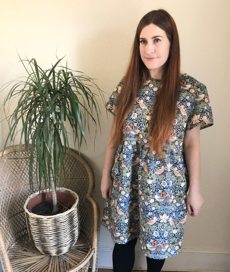May include: A woman wearing a blue and green floral dress with short sleeves. The dress has a pattern of birds and flowers. She is standing in front of a potted plant.
