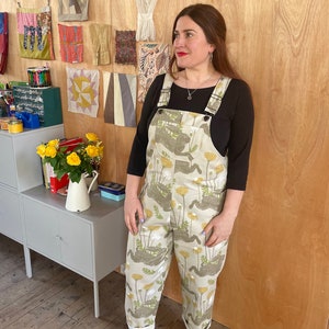 May include: A woman wearing a pair of beige overalls with a floral print. The overalls have a black top and adjustable straps. The woman is standing in front of a wall with a variety of fabric samples.