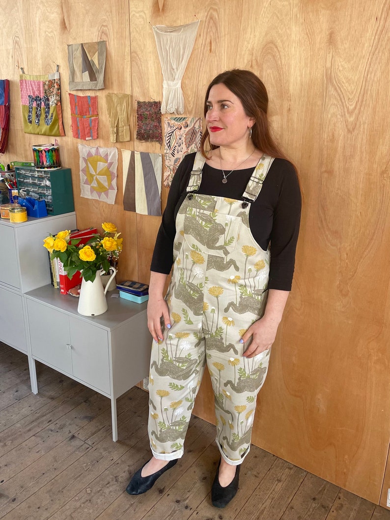 May include: A woman wearing a pair of beige overalls with a floral print. The overalls have a black top and adjustable straps. The woman is standing in front of a wall with a variety of fabric samples.