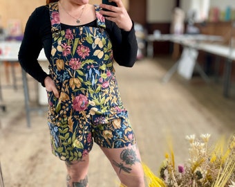 May include: A person wearing a dark blue floral print overall shorts with a black long-sleeved shirt. The person is standing in a room with a bouquet of dried flowers in the foreground.