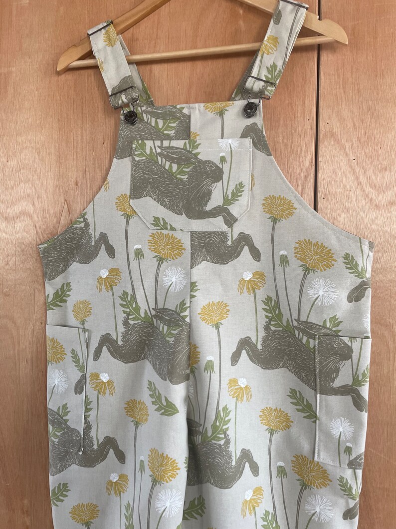 May include: Beige overalls with a pattern of brown rabbits and yellow dandelions. The overalls have adjustable straps and two pockets.