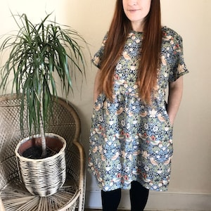 May include: A woman wearing a blue and white floral print dress with short sleeves and a gathered waist. She is standing in front of a wicker chair with a potted plant.