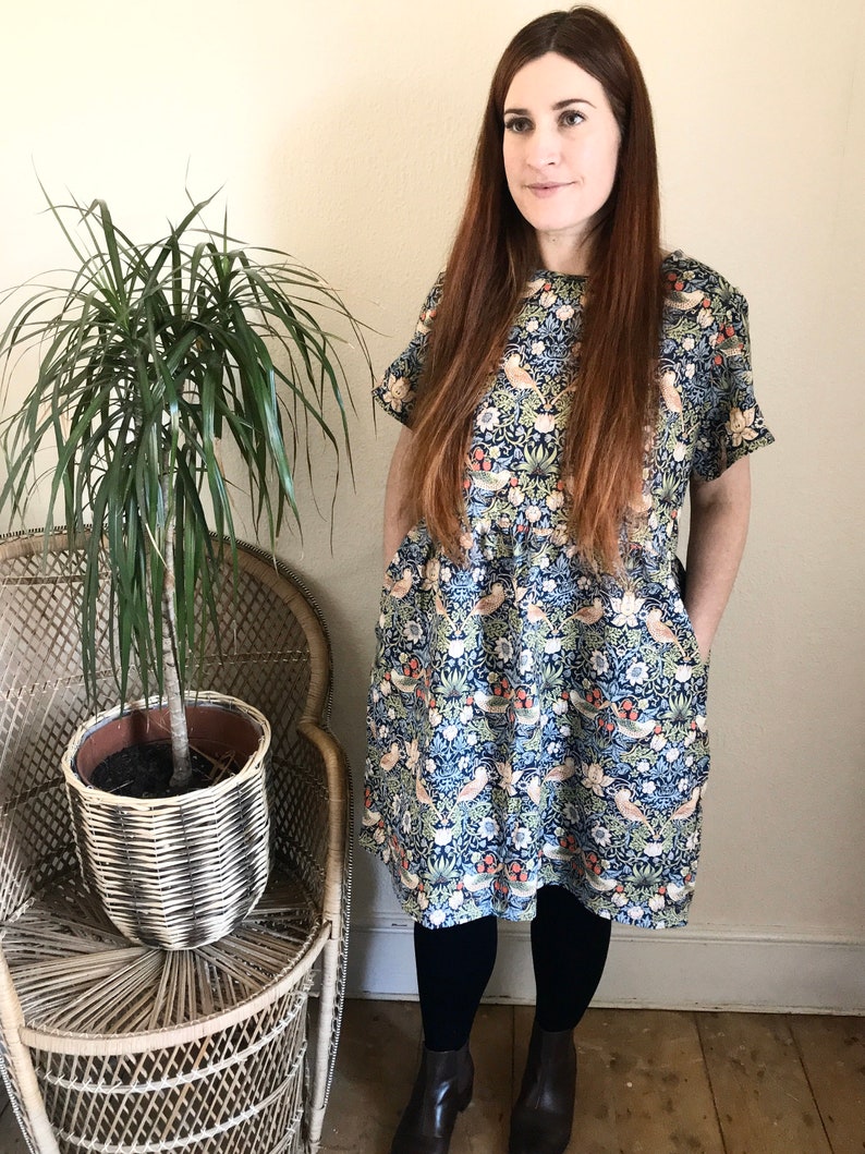 May include: A woman wearing a blue and white floral print dress with short sleeves and a gathered waist. She is standing in front of a wicker chair with a potted plant.