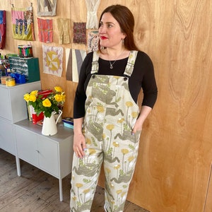 May include: A person wearing a pair of light beige overalls with a dandelion print. The overalls have adjustable straps and pockets. The person is standing in front of a wooden wall with fabric samples hanging on it.