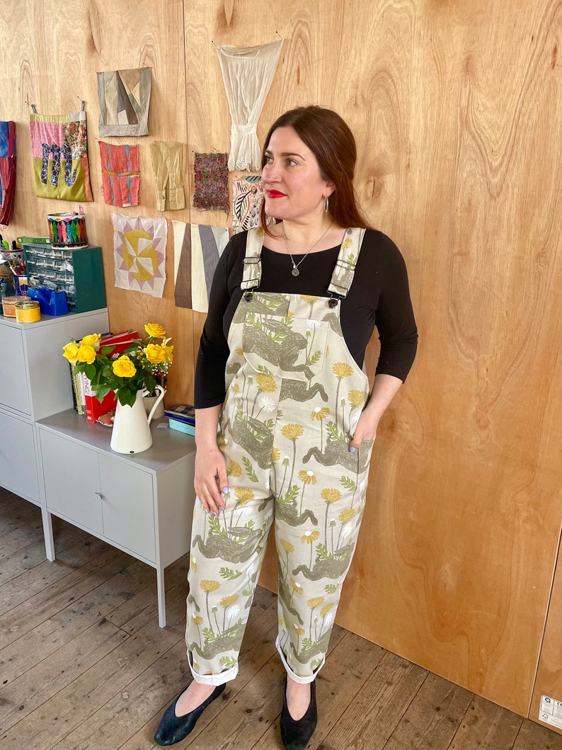 May include: A person wearing a pair of light beige overalls with a dandelion print. The overalls have adjustable straps and pockets. The person is standing in front of a wooden wall with fabric samples hanging on it.