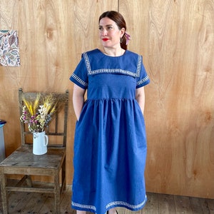 May include: A blue midi dress with a white and blue gingham trim. The dress has a sailor collar and short sleeves. The model is wearing black flats.