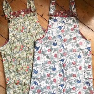 May include: Two pairs of denim overalls with floral patterns. One pair is green with yellow flowers and the other is white with blue birds and red flowers.