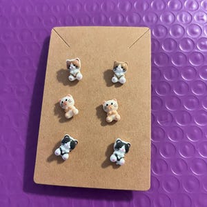 May include: Six cat-shaped stud earrings on a brown card. The earrings are in various colors, including white, brown, and black. The background is a textured purple surface. These earrings are a cute accessory.