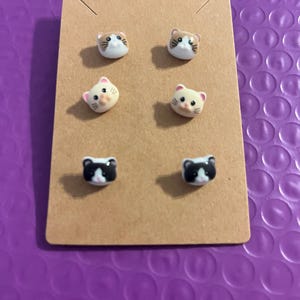 May include: Six pairs of cat-shaped stud earrings on a brown cardboard display. The earrings feature various cat face designs in white, tan, and black. The background is a textured purple surface.