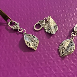 May include: Silver-toned leaf charms with heart-shaped clasps. The charms have detailed leaf patterns and are displayed on a purple textured surface. One charm is attached to a zipper, while two others are separate.