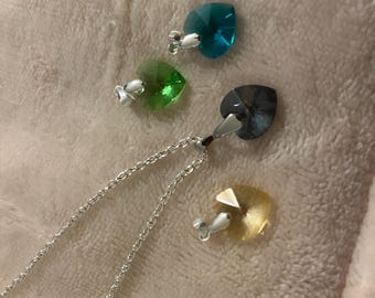 Pode incluir: Um colar de corrente prateada com quatro pingentes em forma de coração nas cores turquesa, verde, cinza e amarelo. Cada pingente tem uma argola prateada. O colar é exibido em uma superfície macia e clara.