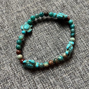 May include: A turquoise and green beaded bracelet with three turtle charms. The bracelet features a mix of round beads in shades of green, brown, and turquoise. The turtle charms are a bright turquoise color.