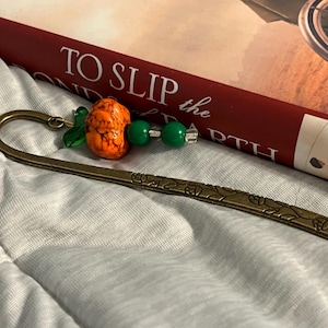 May include: A decorative bookmark with a bronze finish and a pumpkin-shaped bead. The bookmark features a detailed floral design and is adorned with green beads and a leaf charm. The pumpkin bead is orange with brown marbling.