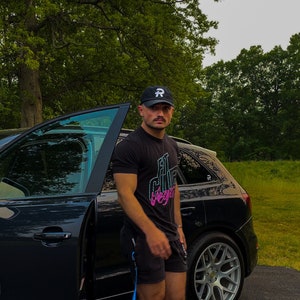 May include: A man wearing a black baseball cap with a white letter 'R' on it, a black t-shirt with a pink and blue graphic that says "Fit Club Vegan", black shorts, and white sneakers. He is standing next to a black car with the door open.