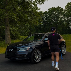 May include: A black SUV with a black man wearing a black shirt and a black hat walking away from the car. The car has a black grill with a silver Audi logo. The car is parked on a black asphalt driveway.