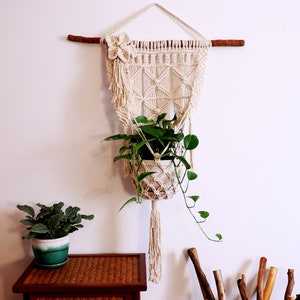 May include: A macrame plant hanger with a white cotton cord and a wooden dowel. The hanger is decorated with a white flower and has a small potted plant hanging from it. The plant is a trailing vine with green leaves.