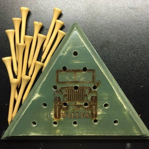 May include: A wooden golf tee holder in the shape of a triangle, painted green with a distressed finish. The holder features a laser-etched image of a vintage Jeep and the word "WILLYS". A bundle of wooden golf tees is next to the holder.