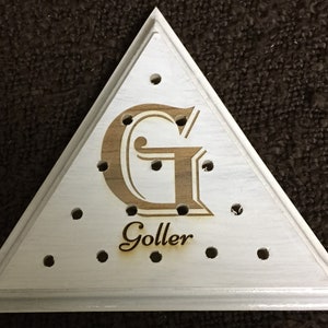 May include: Triangular wooden sign with a white painted surface and a decorative border. The sign features a large, engraved letter "G" in a light brown color, along with the name "Goller" below. Small holes are evenly spaced around the edges.